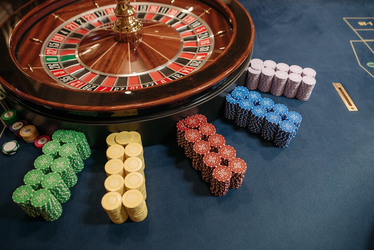 gallery-6 Vibrant poker chips stacked beside a roulette wheel in a casino setting.